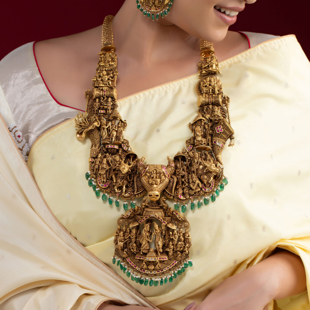 Traditional South Indian temple jewellery necklace with intricate deity motifs, emerald bead drops and gemstone accents, styled with an ivory silk saree.