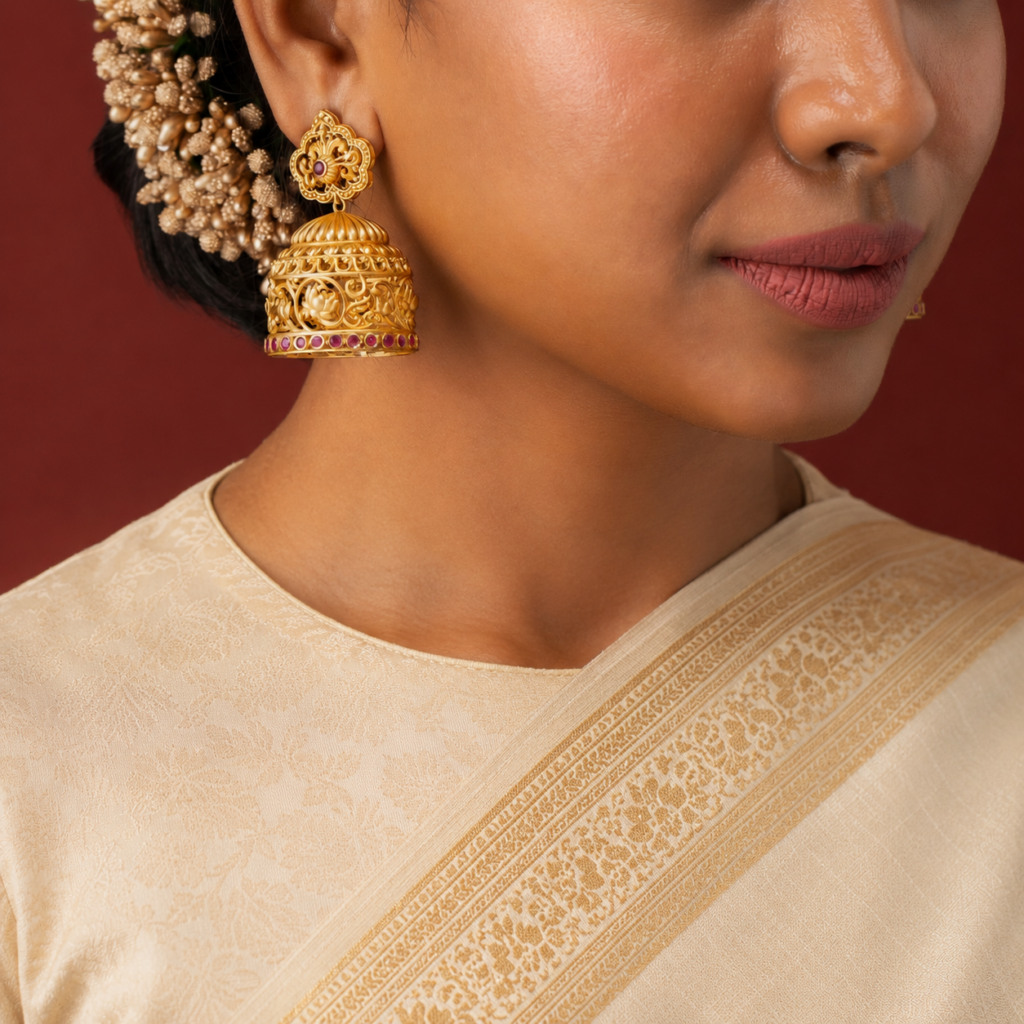 Traditional South Indian gold jhumka earrings featuring intricate floral openwork, sculpted motifs and ruby accents, styled with an ivory silk saree.